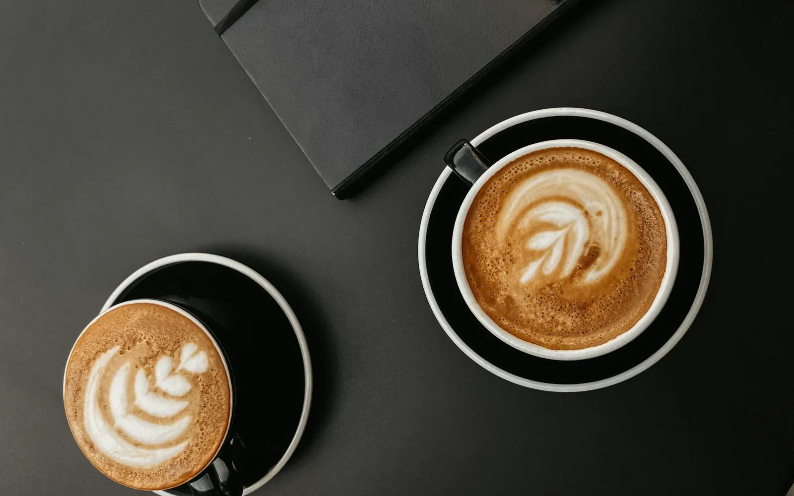 Zwei Cappuccino-Tassen und ein Notizbuch auf einem dunklen Tisch – Sie sucht Ihn Anzeigen im Vergleich